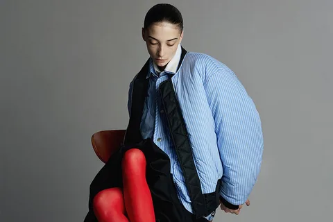 Personne en veste matelassée bleue et collants rouges, assise, regard vers le bas.