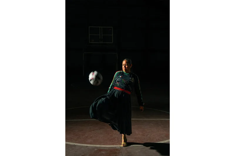 Femme jouant au football dans une robe traditionnelle, éclairage dramatique.