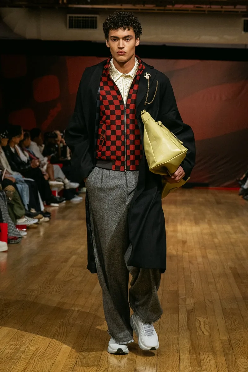 Homme défilant avec un manteau, un sac jaune et un pull à carreaux rouges et noirs.