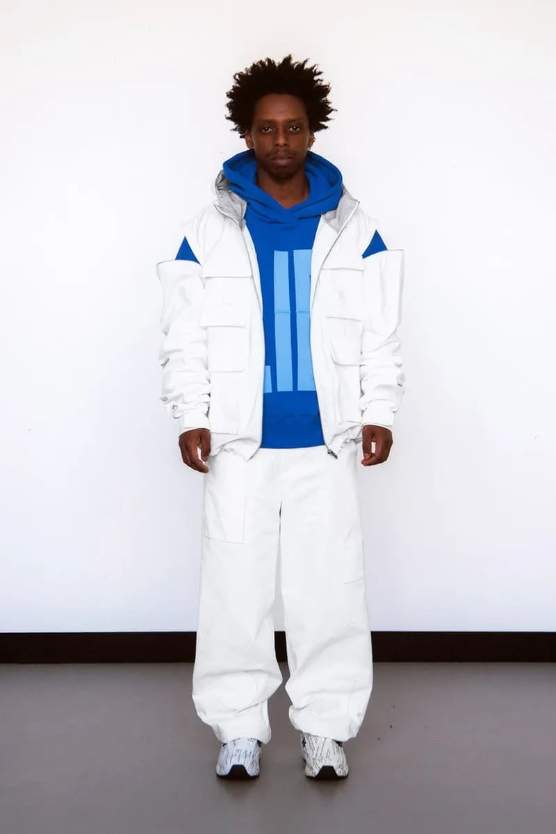 Homme en tenue blanche et bleue, pose devant un mur blanc.