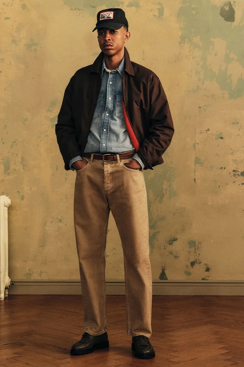 Homme en veste marron, chemise en jean et pantalon beige, devant un mur patiné.
