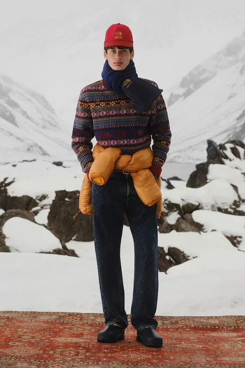 Homme en tenue d'hiver colorée avec bonnet rouge et écharpe, fond montagneux enneigé.
