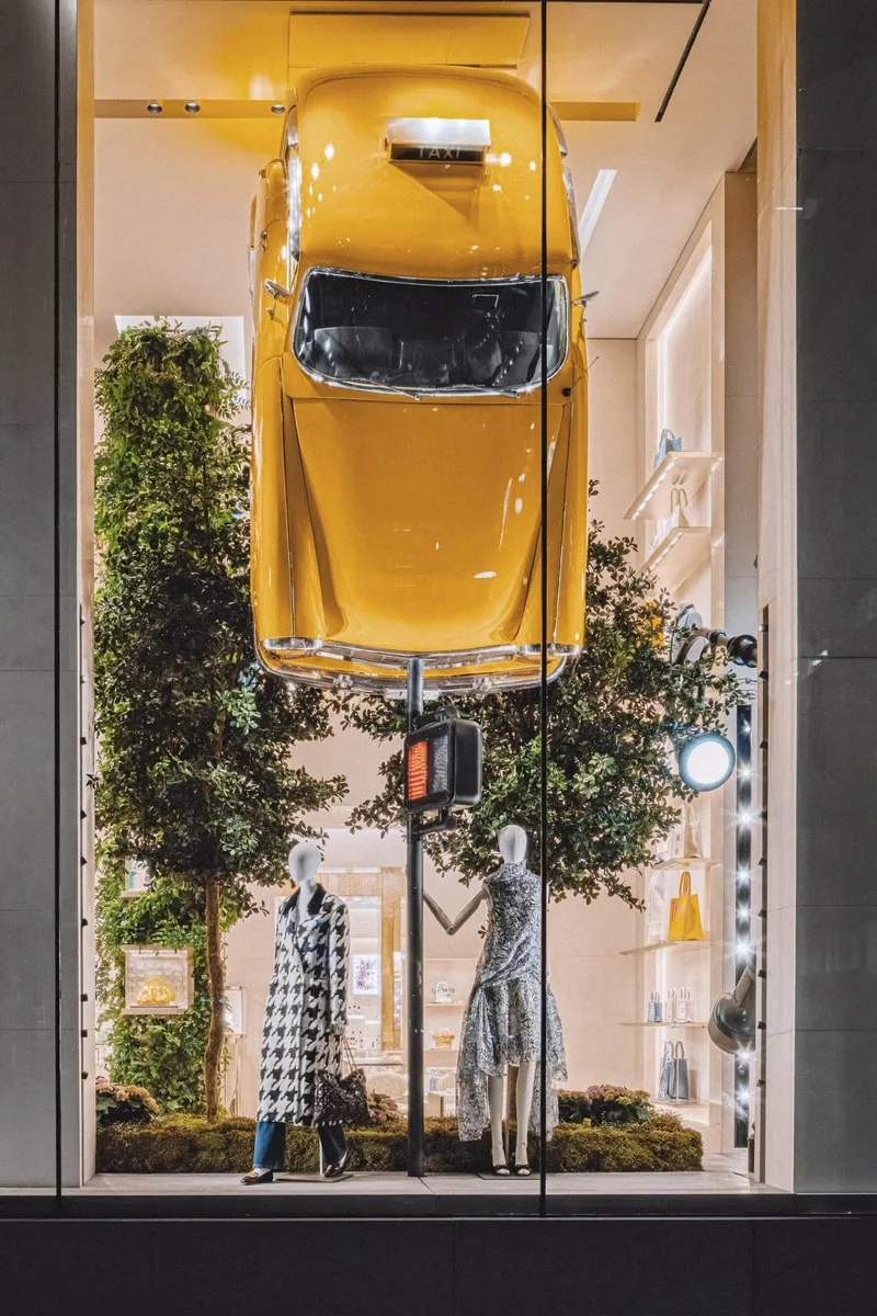 Vitrine avec mannequin et taxi jaune suspendu, arbres en arrière-plan.