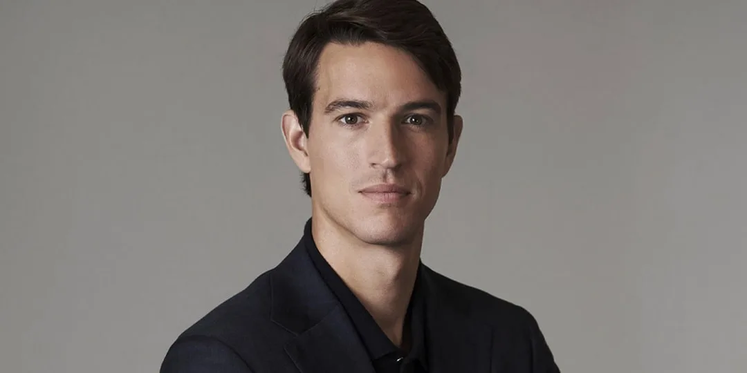 Homme en costume sombre, regard sérieux, fond gris neutre.