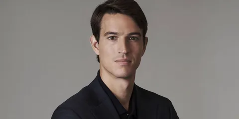 Homme en costume sombre, regard sérieux, fond gris neutre.