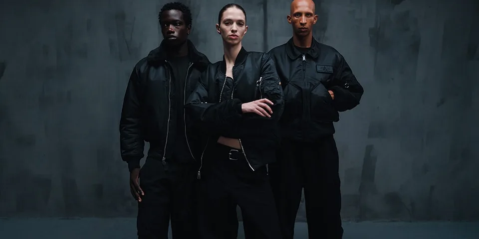 Trois mannequins en vêtements noirs posent devant un fond sombre.