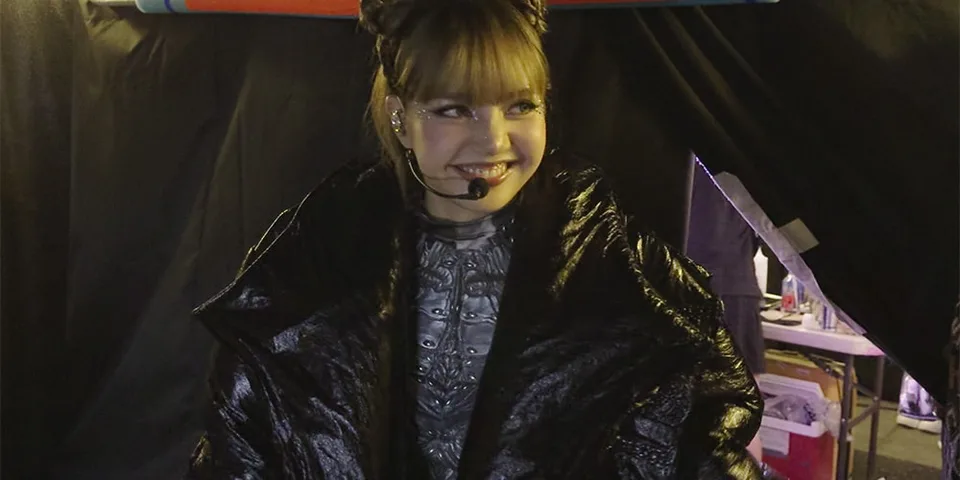 Femme avec veste noire et micro, souriante devant un rideau noir.