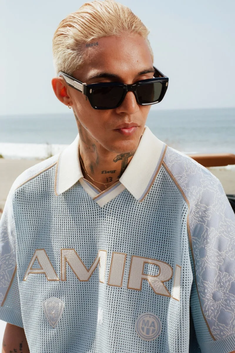 Homme blond portant des lunettes de soleil et un maillot AMIRI près de la plage.