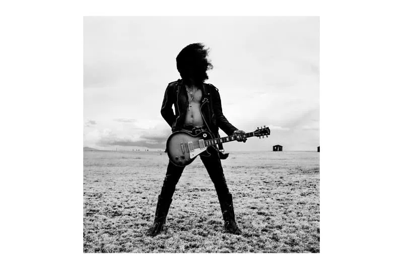 Personne avec une guitare électrique dans un champ, cheveux devant le visage.