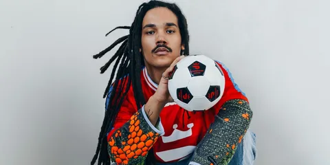 Personne avec des dreadlocks et un ballon de football, portant un maillot multicolore.