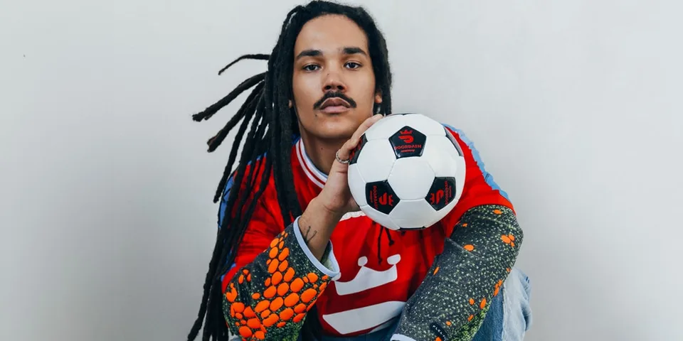 Personne avec des dreadlocks et un ballon de football, portant un maillot multicolore.