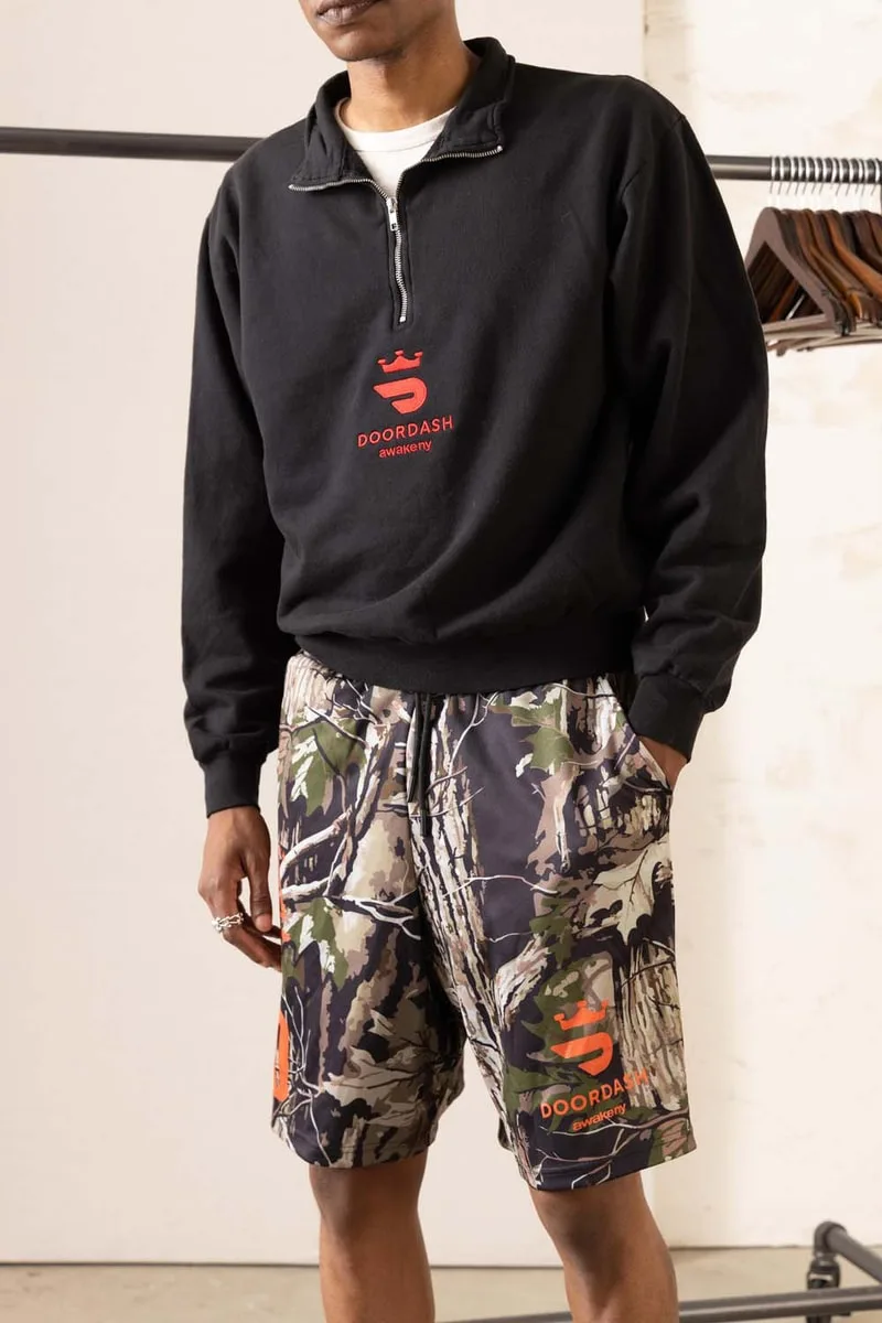 Personne en sweat-shirt noir et short camouflage avec logo rouge.
