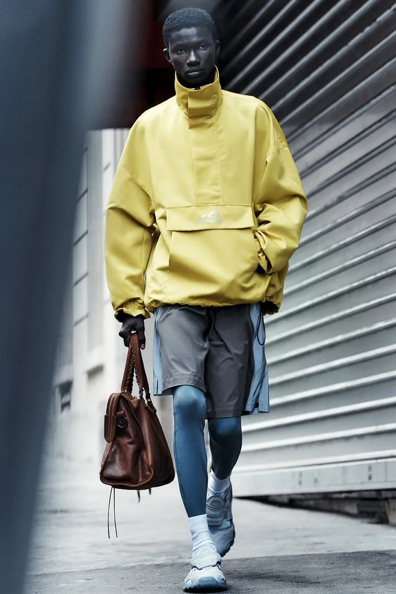 Homme en manteau jaune, short gris, collants bleus, sac brun, baskets blanches.