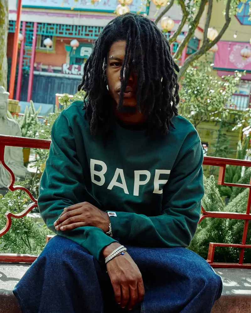 Homme avec sweat BAPE vert, cheveux locks, assis devant décor asiatique.