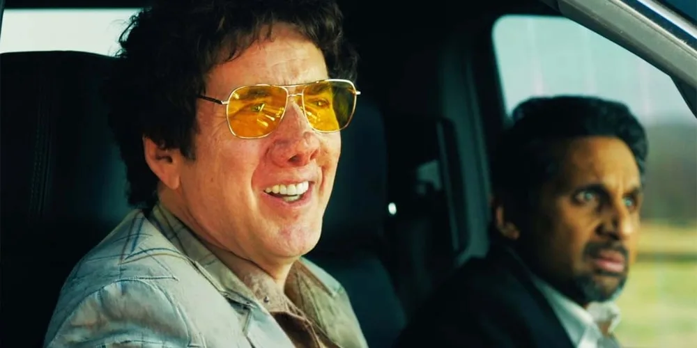 Deux hommes sont assis dans une voiture, l’un portant des lunettes jaunes.