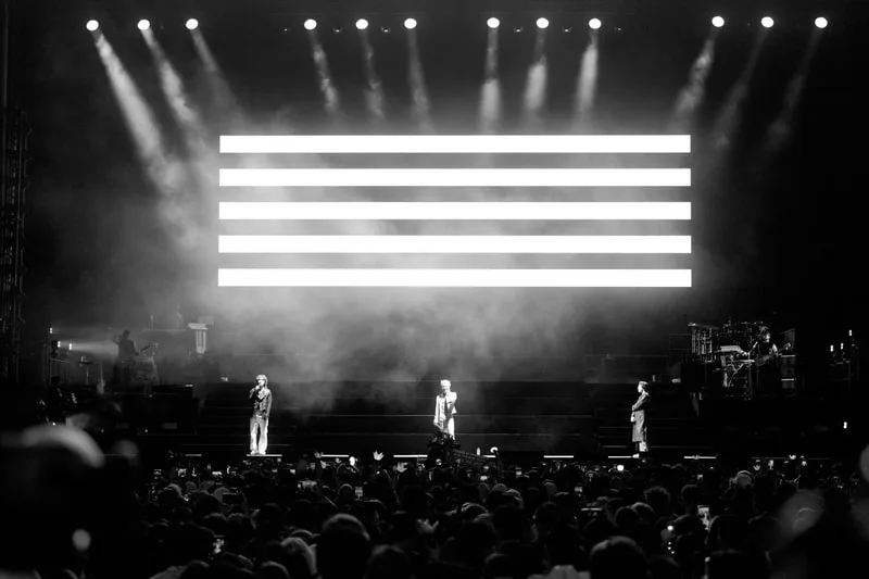 Concert en noir et blanc avec trois artistes sur scène et un grand écran lumineux.