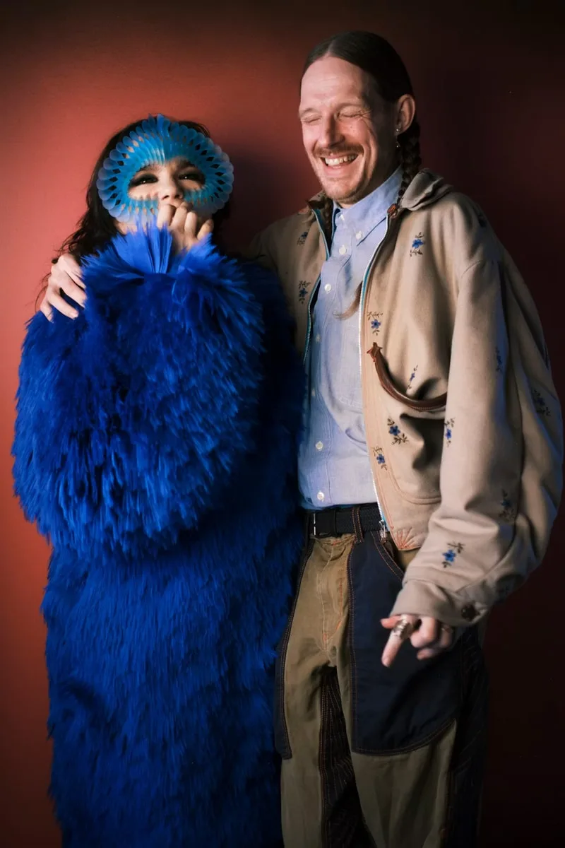 Deux personnes souriantes, l'une en bleu avec masque, l'autre en tenue beige.