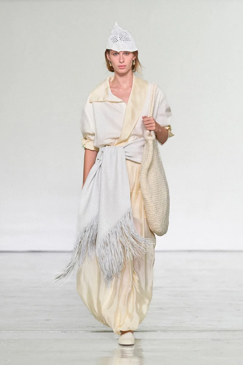 Un modèle défile avec une tenue crème et un grand sac.