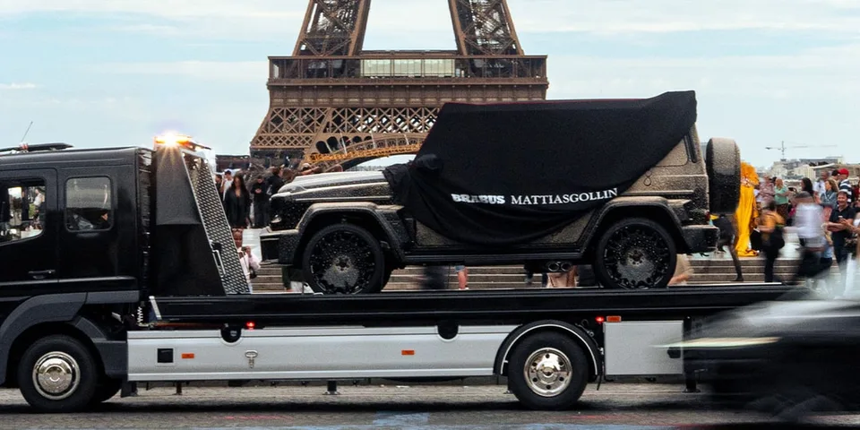 Voiture couverte sur un camion-plateau devant la tour Eiffel, foule en arrière-plan.