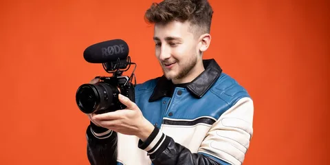 Un homme tenant une caméra avec un micro, fond orange.