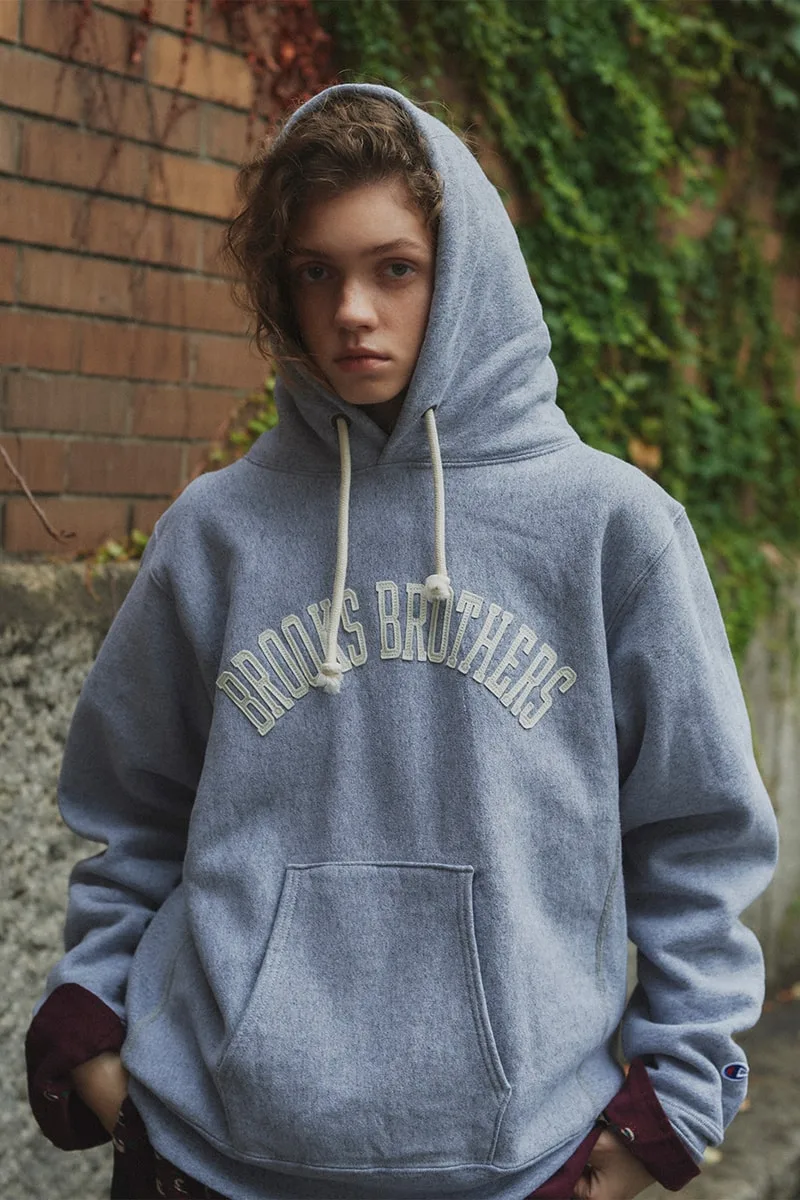 Personne portant un sweat à capuche Brooks Brothers gris devant un mur de briques.