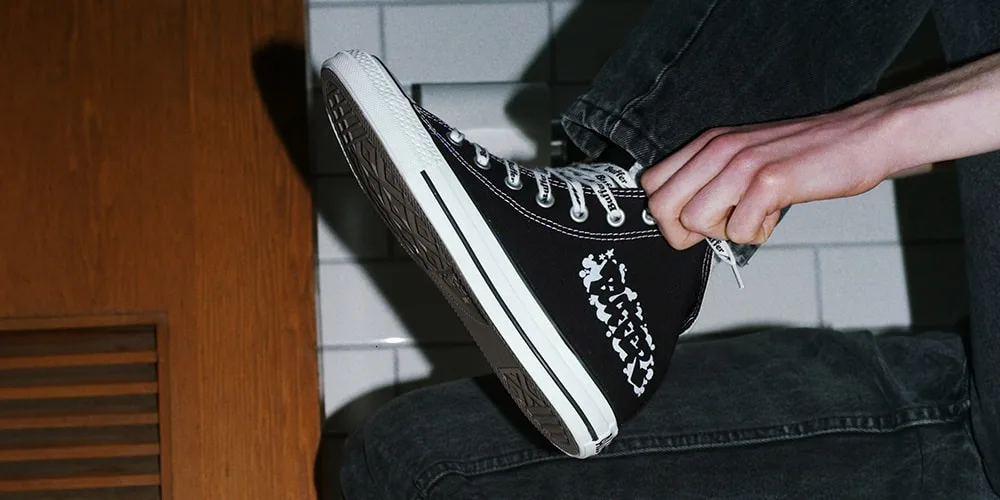 Personne portant des baskets noires avec motif blanc, sur fond de carrelage.