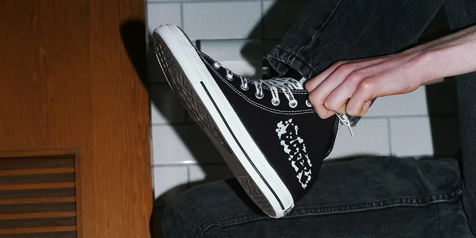 Personne portant des baskets noires avec motif blanc, sur fond de carrelage.