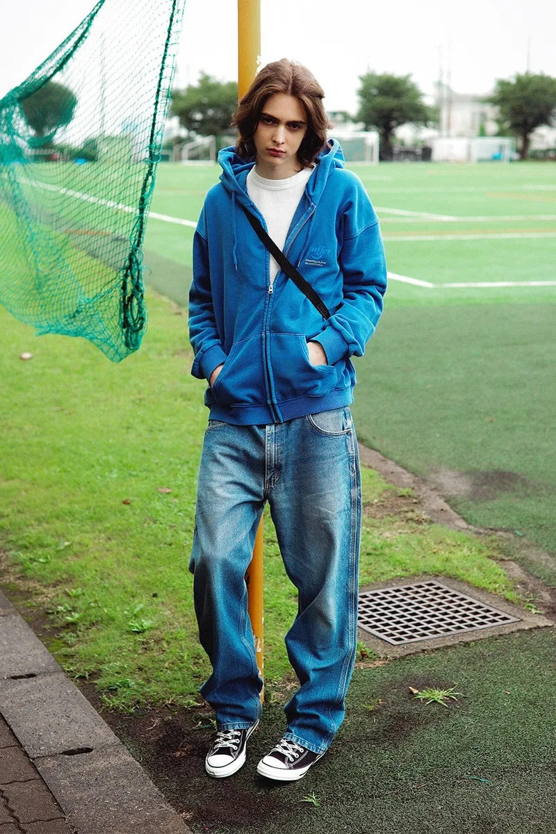 Homme en sweat bleu et jeans, appuyé sur un poteau près d'un terrain de sport.