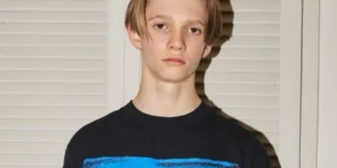 Jeune homme aux cheveux courts devant des volets clairs.