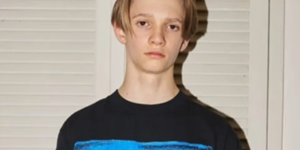 Jeune homme aux cheveux courts devant des volets clairs.