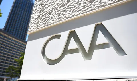 Logo CAA sur mur blanc avec immeubles en fond.