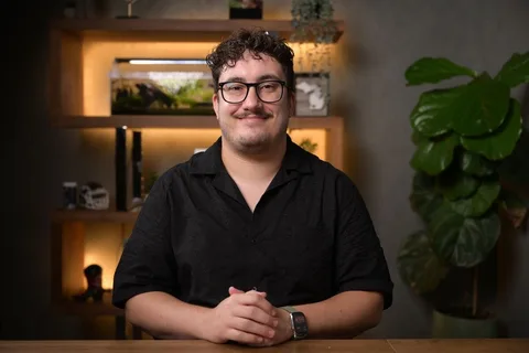 Homme assis à une table, fond avec plante et étagères éclairées.