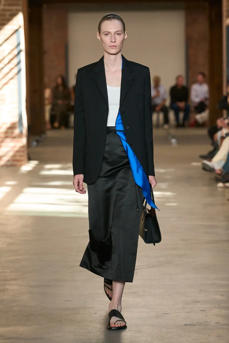 Femme en manteau noir avec jupe satinée, défilant sur un podium.