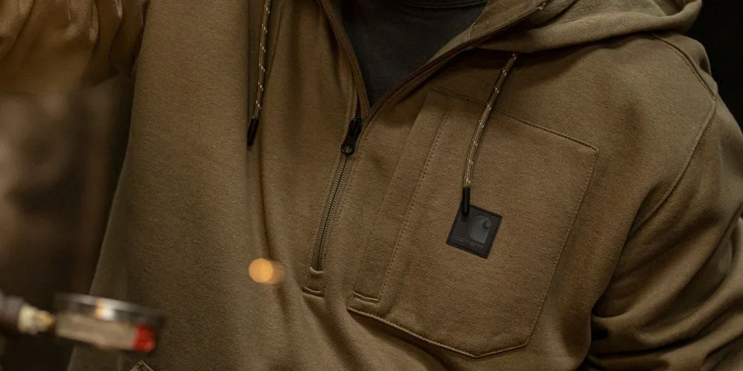 Personne portant un sweat à capuche kaki avec une poche zippée.
