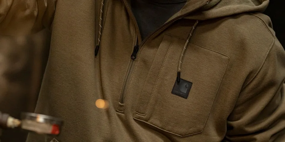 Personne portant un sweat à capuche kaki avec une poche zippée.