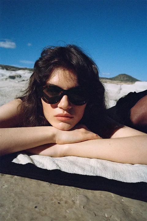 Femme allongée sur une serviette à la plage avec des lunettes de soleil, ciel bleu.