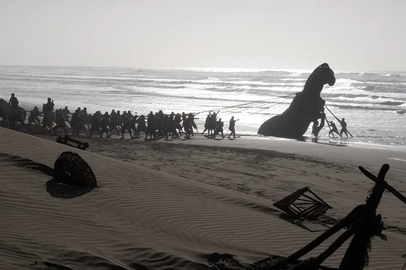 Personnes tirant une grande statue sur une plage, ciel et mer en arrière-plan.