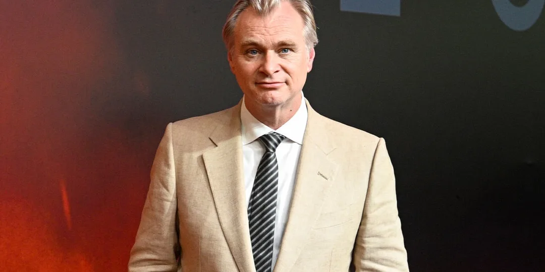 Homme en costume beige avec cravate noire sur fond sombre.