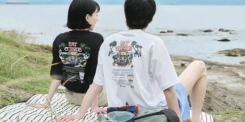 Deux personnes assises près d'un lac avec des t-shirts 'EAT OUTSIDE'.