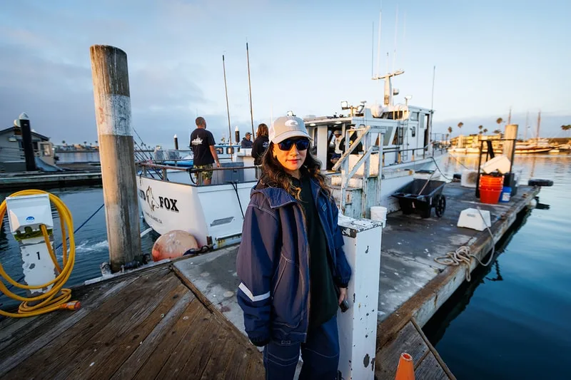 Femme en tenue d'extérieur sur un quai avec bateau au coucher du soleil.