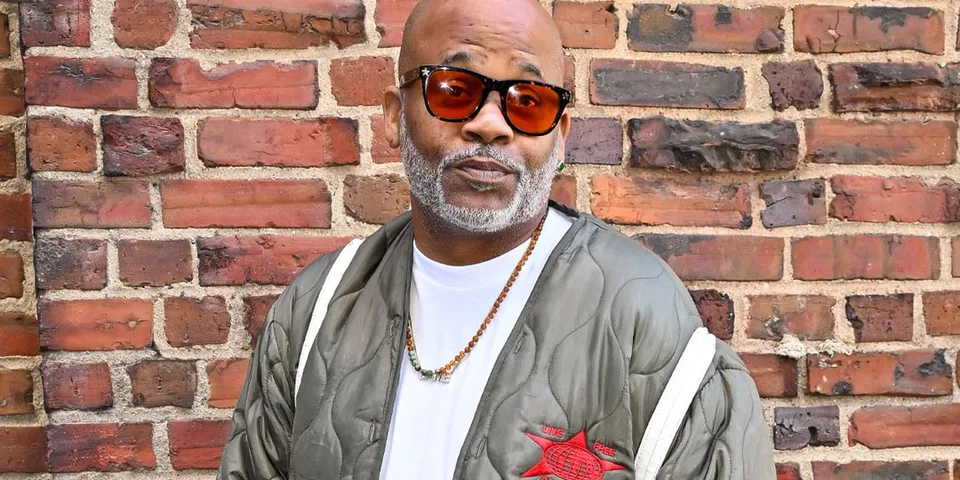 Homme portant des lunettes orange et une veste grise devant un mur de briques.