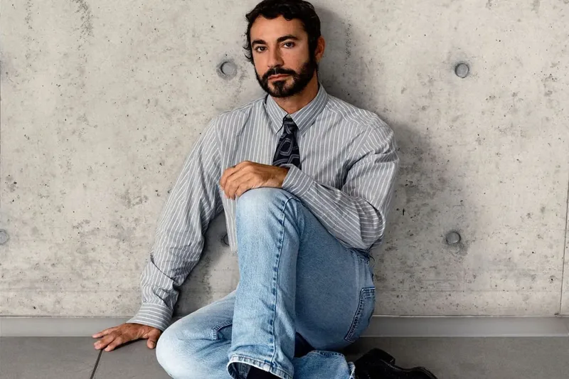 Homme en chemise rayée assis, fond de mur de béton.
