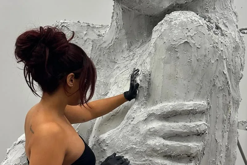 Une femme sculpte une grande statue texturée en argile grise.