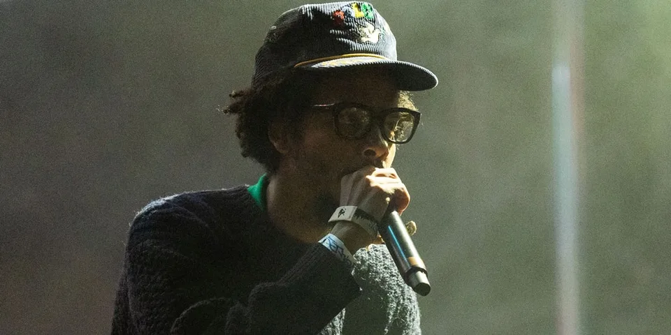 Un homme chante sur scène avec un micro, portant chapeau et lunettes.