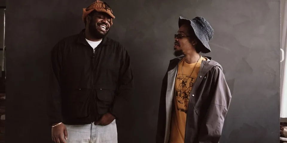 Deux hommes souriants portant des chapeaux et vestes devant un fond sombre.