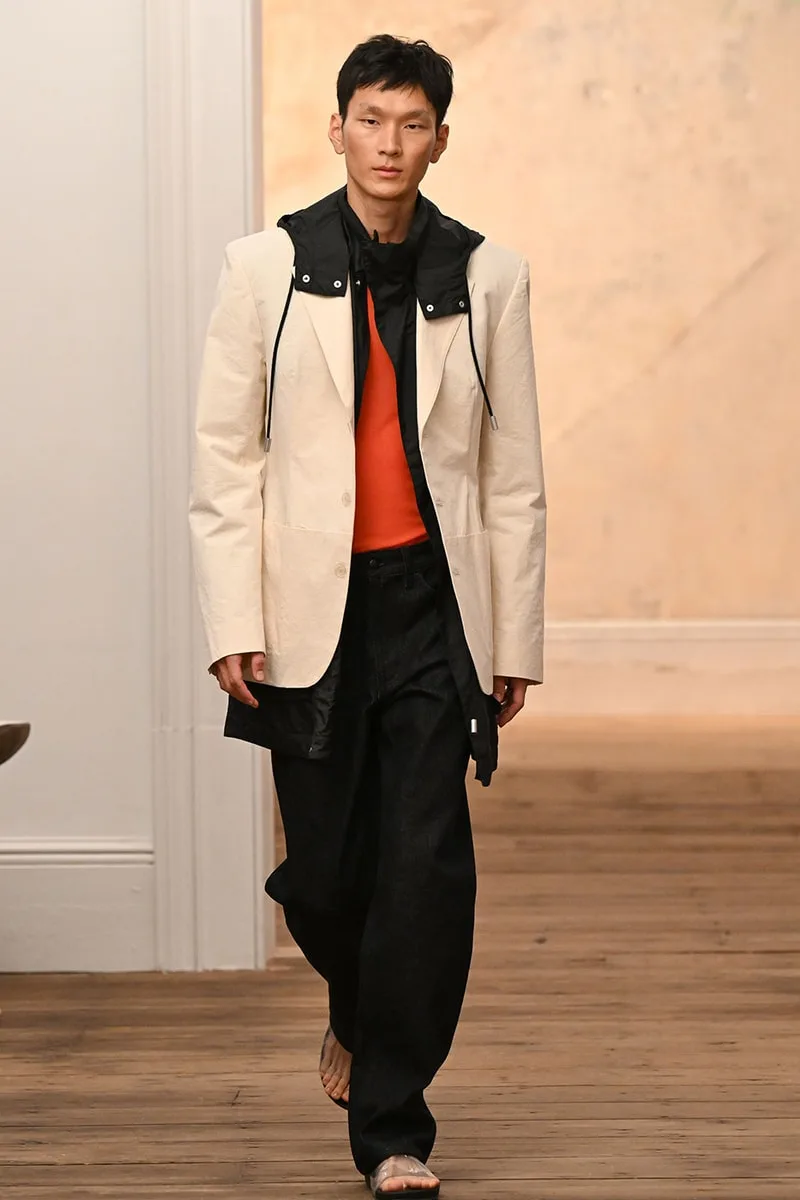 Homme en blazer blanc et pantalon noir sur un podium en bois.