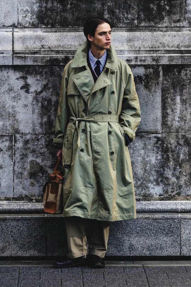 Homme en trench-coat beige avec une mallette devant un mur en pierre.