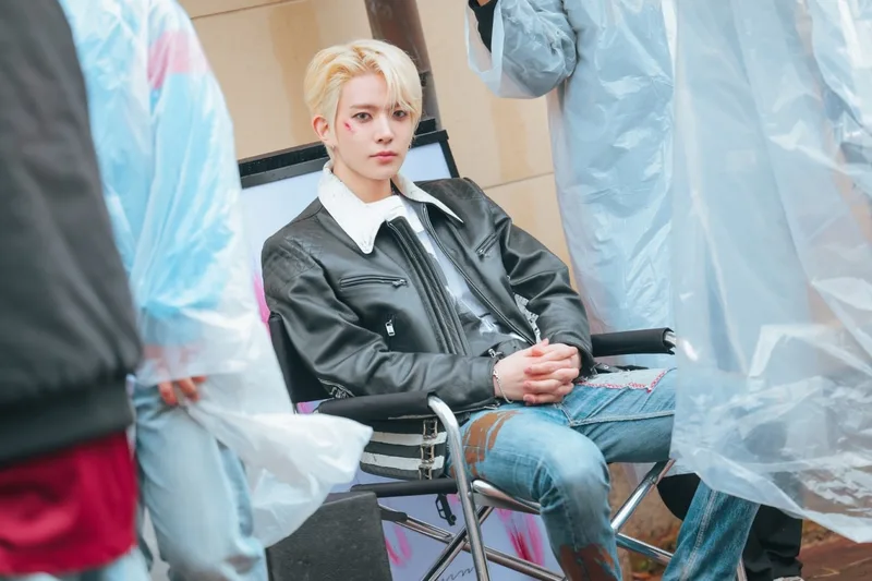 Jeune homme assis sur une chaise, veste en cuir, entouré de personnes en plastique transparent.