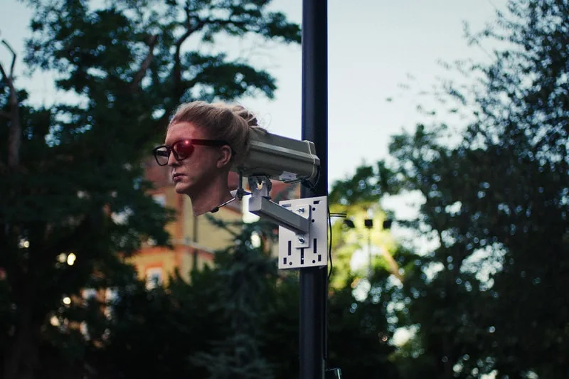 Caméra de surveillance avec tête humaine et lunettes sur poteau.