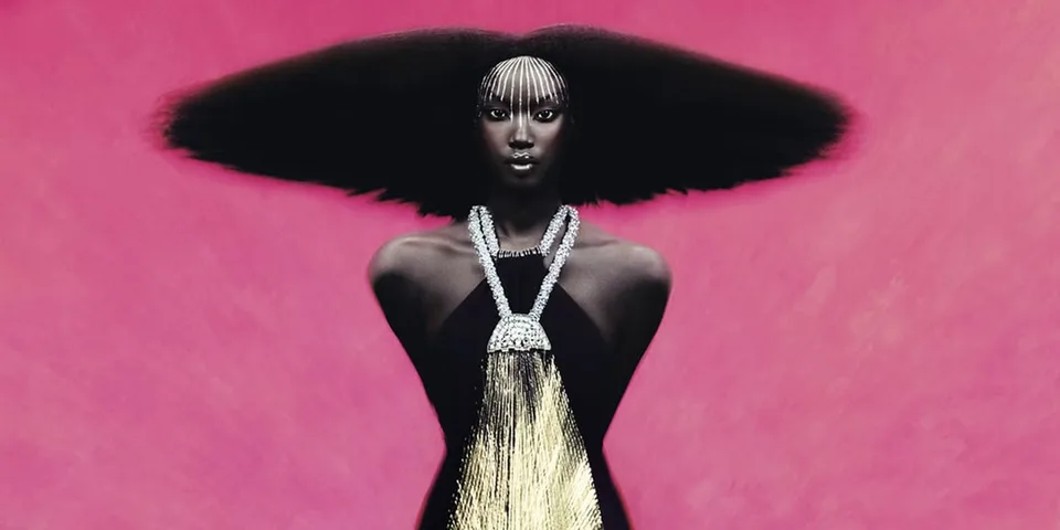 Mannequin à la coiffure afro spectaculaire et au collier doré sur fond rose.